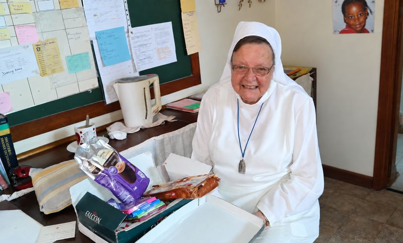 Sister Dolores smiling, seated in her white habit beside a table and noticeboard.